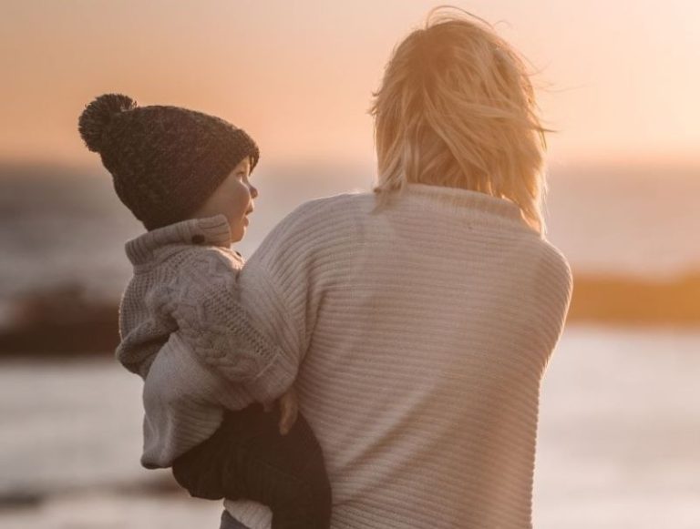 Maman trop inquiète, vue de dos, portant son enfant devant un coucher de soleil, symbole de lien affectif et de sérénité maternelle