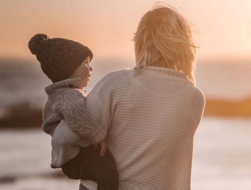 Maman trop inquiète, vue de dos, portant son enfant devant un coucher de soleil, symbole de lien affectif et de sérénité maternelle