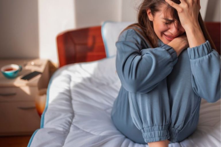 maman qui fait un burn out maternel. Elle craque et pleure assise sur son lit. elle se tient le visage avec les mains.