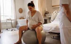 Photo naturelle et chaleureuse d’une femme enceinte installée sur un ballon de grossesse dans une salle de naissance baignée de lumière. L’atmosphère apaisante, aux tons beige et crème, illustre la préparation du projet de naissance et l’accompagnement bienveillant autour de l’accouchement. Le style documentaire et authentique met en avant la sérénité, la confiance et la maternité au naturel.