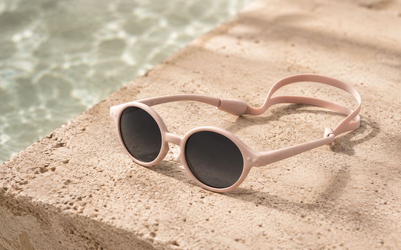 Lunettes de soleil bébé roses avec cordon posées sur une margelle de piscine beige avec eau turquoise en arrière-plan.