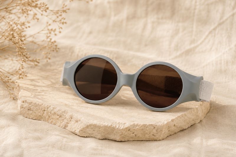 Lunettes de soleil bébé grises avec verres foncés posées sur un fond beige texturé dans une ambiance naturelle et minimaliste.