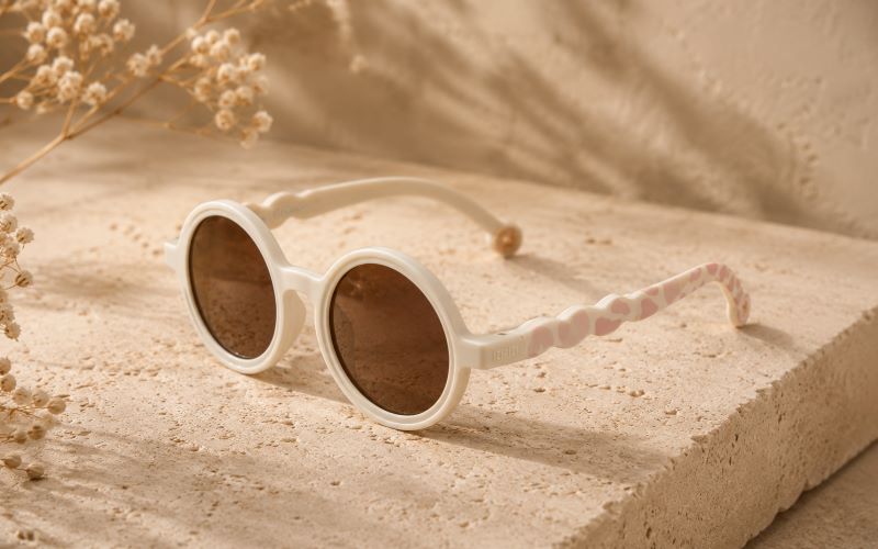 Lunettes de soleil bébé rondes crème avec branches roses posées sur un fond beige texturé et lumineux.