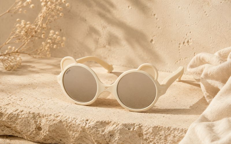 Lunettes de soleil bébé beige avec forme ourson posées sur un fond texturé naturel aux tons sable.