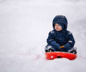 Comment habiller bébé au ski : jeune enfant en combinaison de ski bleu marine assis sur une luge rouge dans la neige.