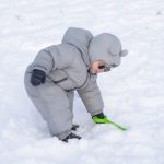 Comment habiller bébé au ski : jeune enfant en combinaison de ski grise et lunettes de soleil jouant dans la neige avec une pelle verte.