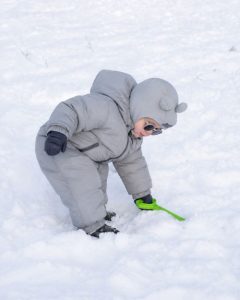 Comment habiller bébé au ski : jeune enfant en combinaison de ski grise et lunettes de soleil jouant dans la neige avec une pelle verte.