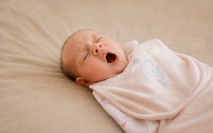 Routine du soir bébé : nouveau-né bâillant paisiblement sur un lit avec un drap-housse beige avant l'endormissement.