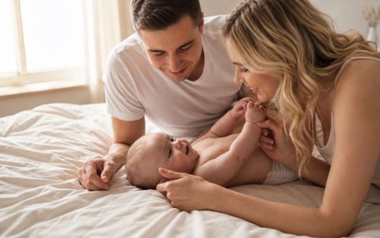 Couple après bébé partageant un moment de tendresse avec leur nourrisson sur un lit dans une chambre lumineuse.