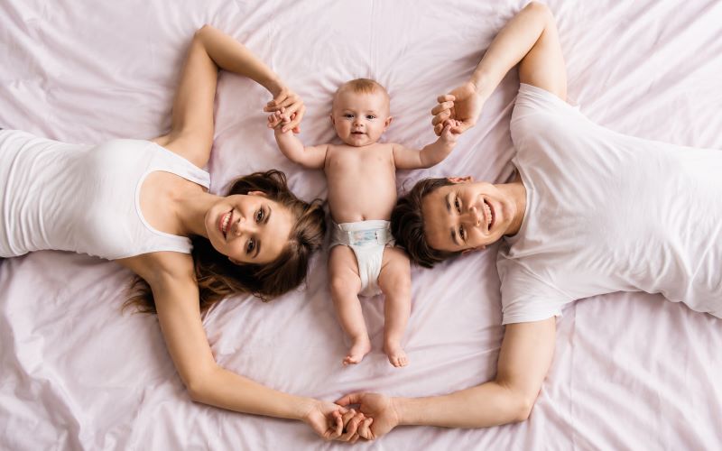 Couple après bébé allongé avec son nourrisson sur un lit, partageant un moment de complicité et de tendresse en famille.